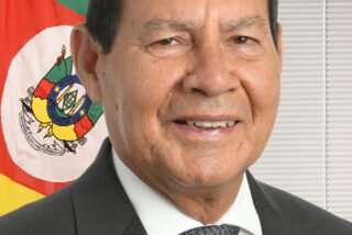 Hamilton Mourão em retrato oficial, de terno e gravata, diante da bandeira do Rio Grande do Sul.