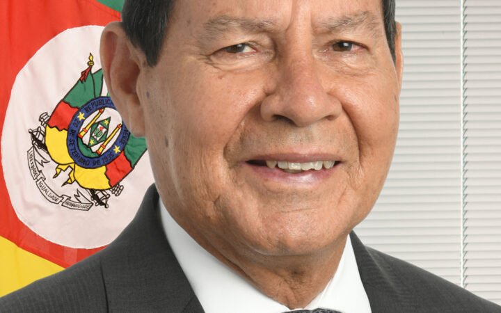 Hamilton Mourão em retrato oficial, de terno e gravata, diante da bandeira do Rio Grande do Sul.